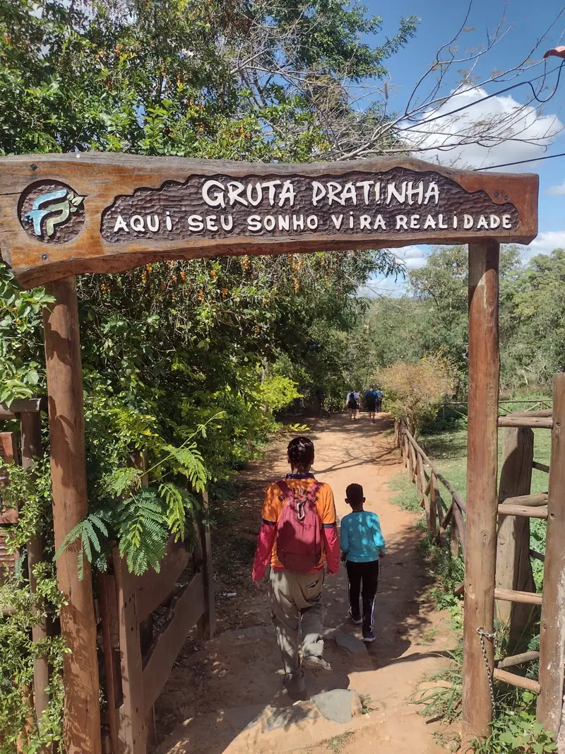 Pórtico de madeira da entrada da Gruta Pratinha, com dois visitantes seguindo pela trilha